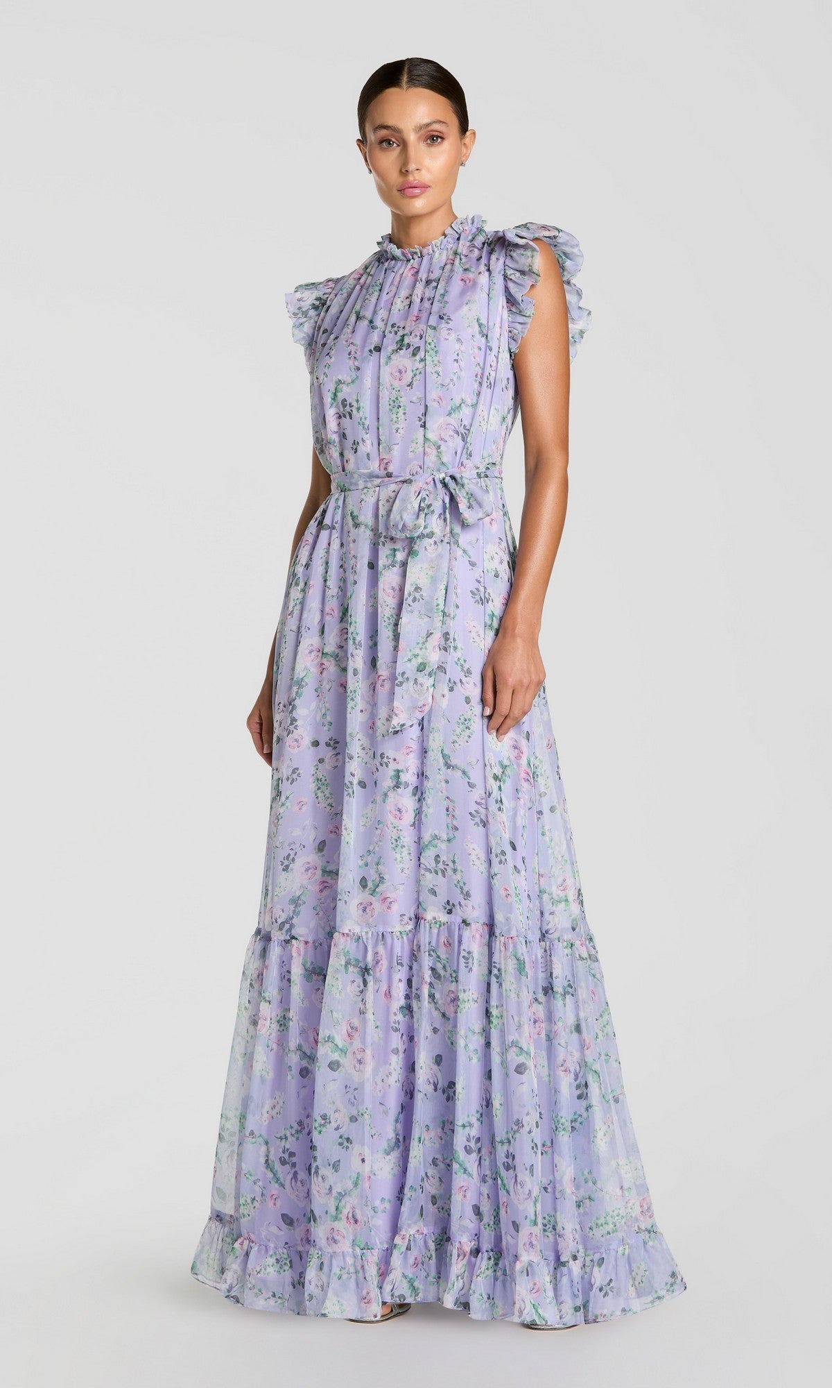 A woman stands against a plain background wearing the Mac Duggal 56116 Lilac Floral-Print Formal Gown, featuring a sleeveless, floor-length design with ruffled shoulders and a waist tie.
